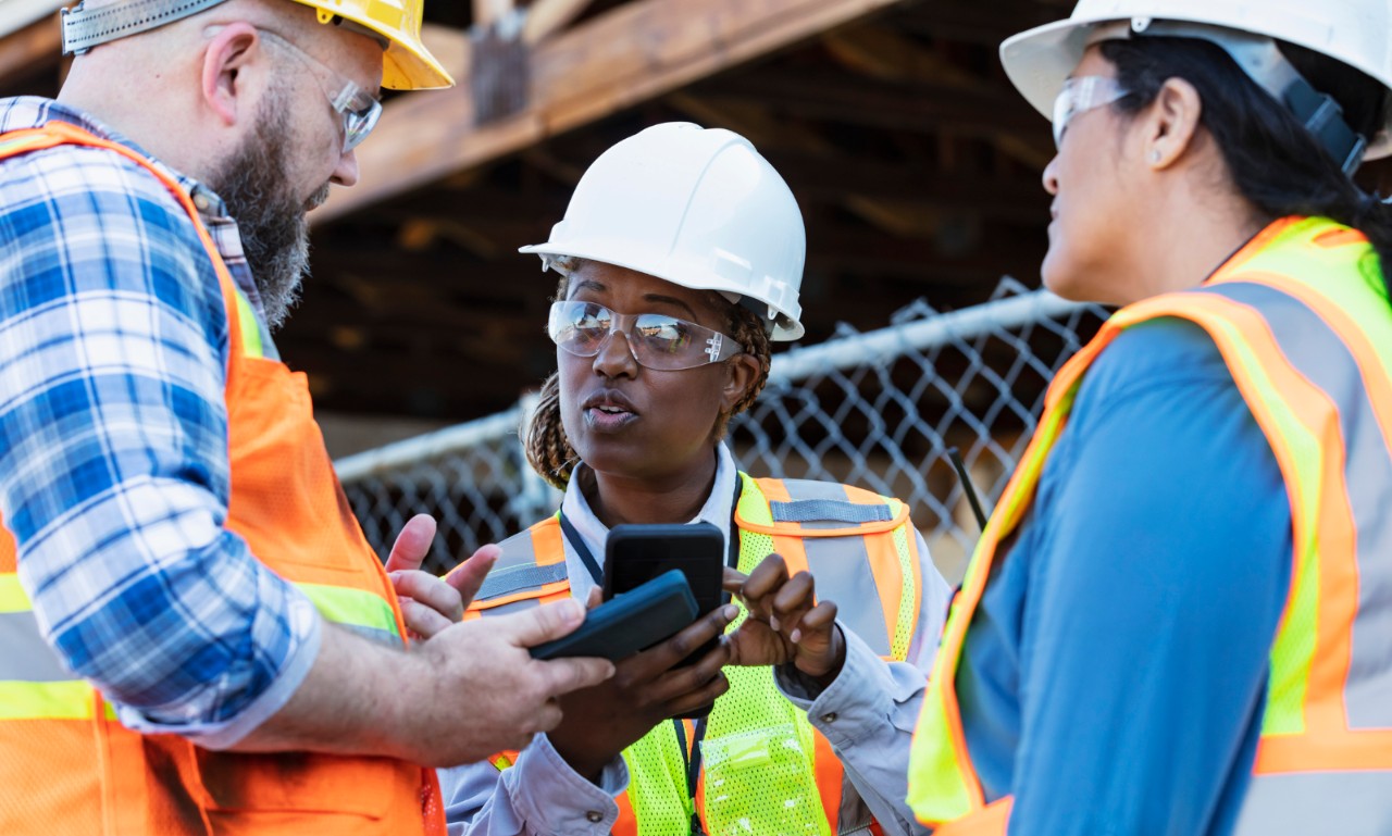 A multiracial group of three construction workers having a meeting at a job site. The focus is on the African-American woman, pointing at her mobile phone, looking at her male coworker with a serious expression. The project is the renovation of a strip mall.